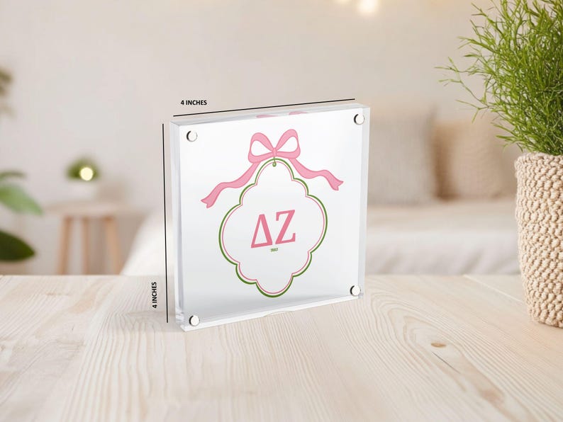 Puede incluir: Un bloque acr&iacute;lico transparente, de aproximadamente 10 cm cuadrados, con un lazo rosa y un emblema decorativo con las letras griegas &Delta;Z en rosa. El emblema est&aacute; delineado en verde y rosa. El bloque se muestra sobre una superficie de madera clara.