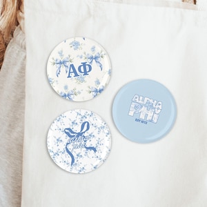 May include: Three round buttons with floral and text designs. One button features the Greek letters Alpha and Phi with blue floral accents. Another has the text "Alpha Phi" and "EST 1872". The third has the text "alpha phi" with a bow.