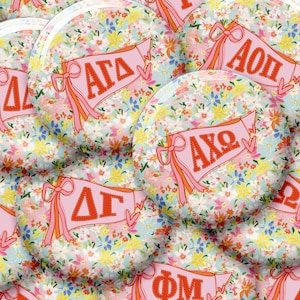 May include: A collection of round buttons with a floral pattern background. Each button features a pink banner with Greek letters and a ribbon design. The buttons have a light blue, yellow, and white floral pattern.