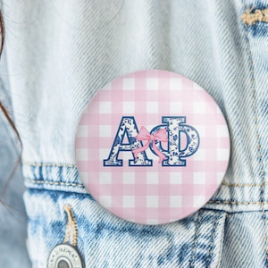 May include: A round button with a pink gingham pattern background. The button features the Greek letters ΑΦ in a blue floral design, with a pink bow. The button is pinned to a denim jacket.
