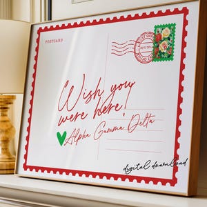 May include: A framed print designed to resemble a postcard. The white background features red text that reads "Wish you were here!" and "Alpha Gamma Delta." A green heart and a floral stamp are also visible. The frame is a light brown color.
