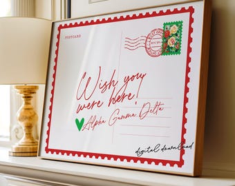 May include: A framed print designed to look like a postcard. The white background has red text saying "Wish you were here!" and "Alpha Gamma Delta." A green heart and a floral stamp are also visible. The frame is light brown.