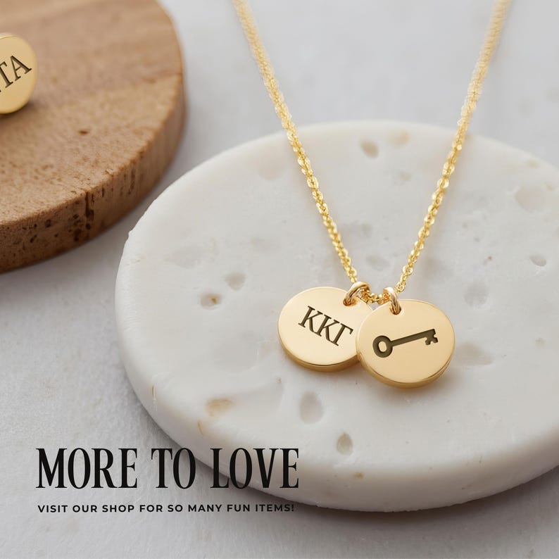 May include: A gold-toned necklace featuring a delicate chain and two circular pendants. One pendant is engraved with the Greek letters KKT, while the other displays a small key icon. The necklace is displayed on a white textured surface.
