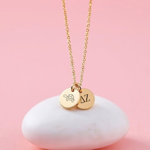 May include: A gold-colored necklace with two circular pendants. One pendant features a turtle design, and the other displays the Greek letters Δ and Z. The necklace is displayed on a white, oval-shaped object against a pink background.