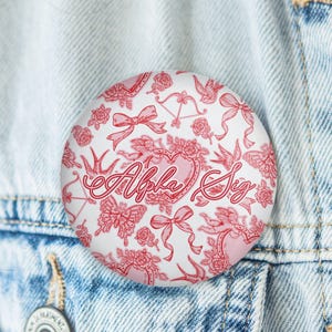 May include: A round, white button with a red floral and bow pattern. The button features the text "Alpha Sig" in a cursive font, surrounded by roses, butterflies, and other decorative elements. The button is pinned to a denim jacket.