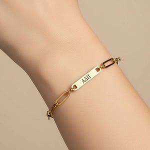 May include: Gold-colored chain bracelet with an oblong plate featuring the engraved letters ΑΔΠ. The bracelet is made of linked oval and rectangular segments. The bracelet is shown on a person's wrist.