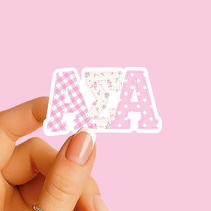 May include: A sticker with the letters ASA in a patchwork design. The letter A is pink gingham, the S is floral, and the other A is pink with white polka dots. The sticker is held against a pink background.