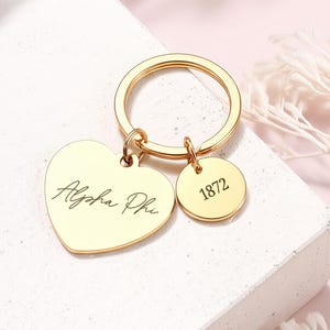 May include: A gold-colored keychain with a heart-shaped charm and a round charm. The heart charm has the script "Alpha Phi" engraved on it. The round charm has the number "1872" engraved on it. The keychain is on a white surface.