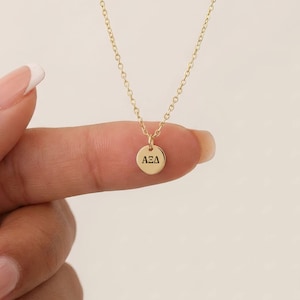 May include: A delicate gold-colored necklace with a small, round pendant. The pendant has the letters AEΔ engraved on it. The necklace is held between fingers against a soft, neutral background. The chain is fine and elegant.