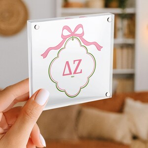 Puede incluir: Un bloque acr&iacute;lico transparente con un dise&ntilde;o de lazo y cinta rosa. El bloque tiene un fondo blanco con un borde decorativo rosa y verde. Las letras "&Delta;Z" se muestran en rosa.