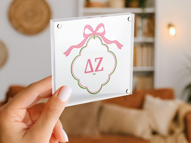 Puede incluir: Un bloque acr&iacute;lico transparente con un dise&ntilde;o de lazo y cinta rosa. El bloque tiene un fondo blanco con un borde decorativo rosa y verde. Las letras "&Delta;Z" se muestran en rosa.