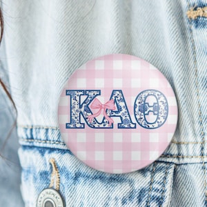 May include: A round button with a pink and white gingham pattern. The letters KAO are displayed in blue with floral designs and a pink bow. The button is pinned to a denim jacket.