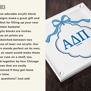 May include: A square acrylic block art sign with a blue ribbon and the Greek letters &Alpha;&Delta;&Pi; in blue. The sign is white with a dark wood grain background. The sign is 4 x 4 inches.