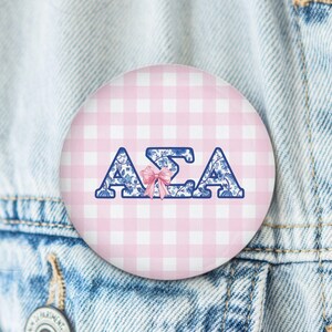 May include: A round button with a pink and white gingham pattern background. The button features the Greek letters ΑΣΑ in blue with a floral design, and a pink bow. The button is pinned to a denim jacket.