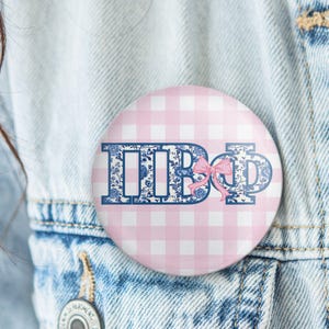 May include: A round button with a pink and white gingham pattern background. The button features the Greek letters IIBΦ in a blue floral design, with a pink bow. The button is pinned to a denim jacket.