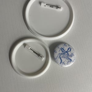 May include: Two large white circular pin-back buttons with metal pin fasteners on the reverse side and one smaller circular button featuring a blue floral pattern with a blue ribbon graphic and the text alpha xi delta.