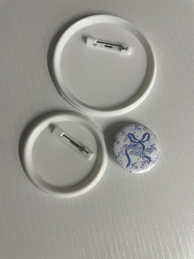 May include: Two large white circular pin-back buttons with metal pin fasteners on the reverse side and one smaller circular button featuring a blue floral pattern with a blue ribbon graphic and the text alpha xi delta.