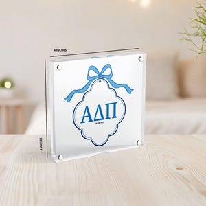 May include: A clear acrylic block, 4 inches square, featuring a blue ribbon and the Greek letters &Alpha;&Delta;&Pi;. The letters are inside a white and blue decorative shape. The text "BE THE FIRST" is below the letters.
