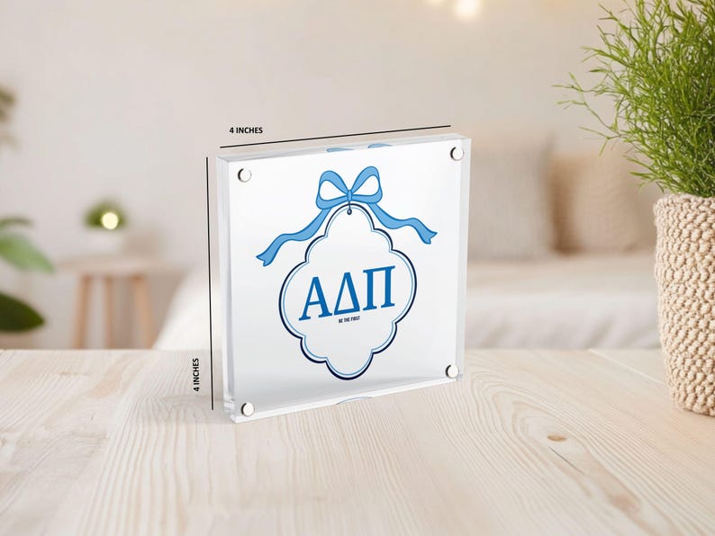 May include: A clear acrylic block, 4 inches square, featuring a blue ribbon and the Greek letters &Alpha;&Delta;&Pi;. The letters are inside a white and blue decorative shape. The text "BE THE FIRST" is below the letters.