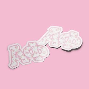 May include: Four stickers with a floral pattern in pink and white. The stickers are in the shape of Greek letters, including Alpha and Phi. The letters have a pink outline and are set against a light pink background.