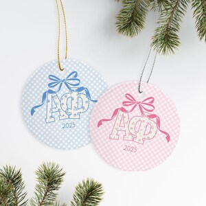 May include: Two round Christmas ornaments. One is light blue with a gingham pattern and a blue bow, the other is pink with a gingham pattern and a pink bow. Both have floral letters and the year 2025.