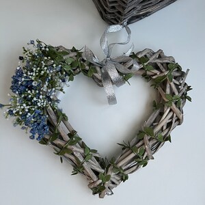 May include: A heart-shaped wreath made of woven twigs and green leaves, decorated with a cluster of blue and white flowers and a silver ribbon.