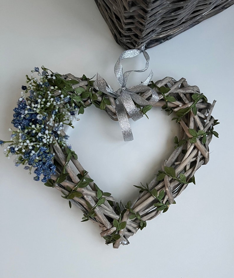 May include: A heart-shaped wreath made of woven twigs and green leaves, decorated with a cluster of blue and white flowers and a silver ribbon.