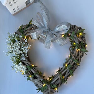 May include: A heart-shaped wicker wreath with a silver ribbon bow and white flowers. The wreath is decorated with small, warm white lights.