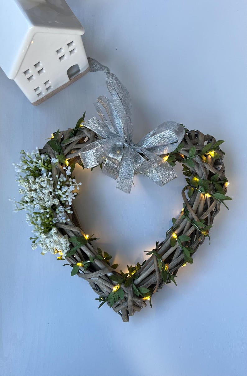 May include: A heart-shaped wicker wreath with a silver ribbon bow and white flowers. The wreath is decorated with small, warm white lights.