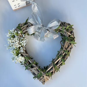 May include: A heart-shaped wreath made of woven twigs and greenery, decorated with white flowers and a silver ribbon bow.