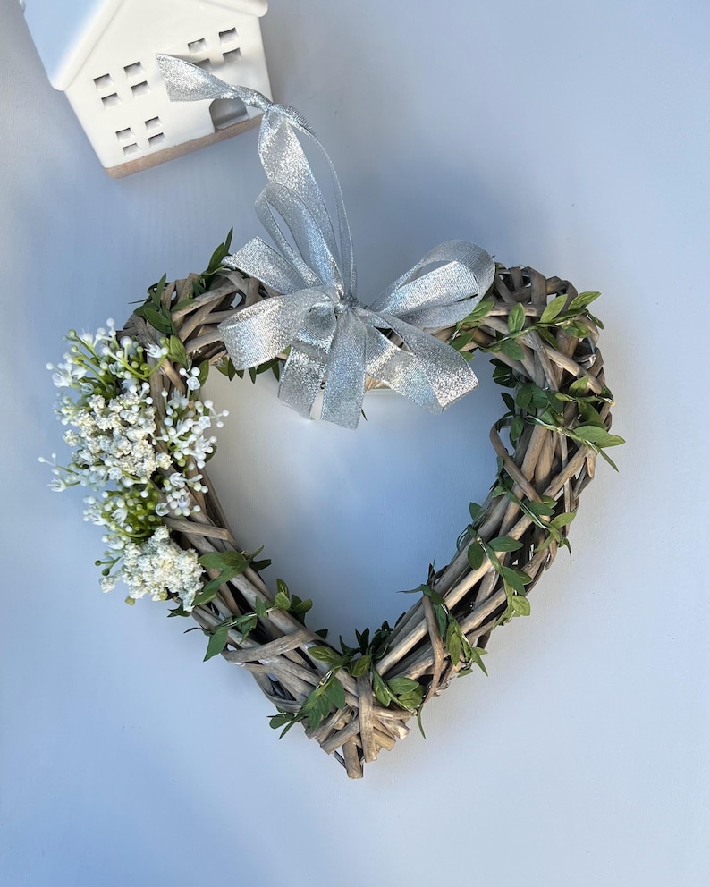 May include: A heart-shaped wreath made of woven twigs and greenery, decorated with white flowers and a silver ribbon bow.