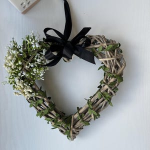 May include: A heart-shaped wreath made of woven twigs and green leaves, decorated with white flowers and a black ribbon.