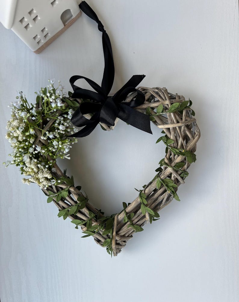 May include: A heart-shaped wreath made of woven twigs and green leaves, decorated with white flowers and a black ribbon.