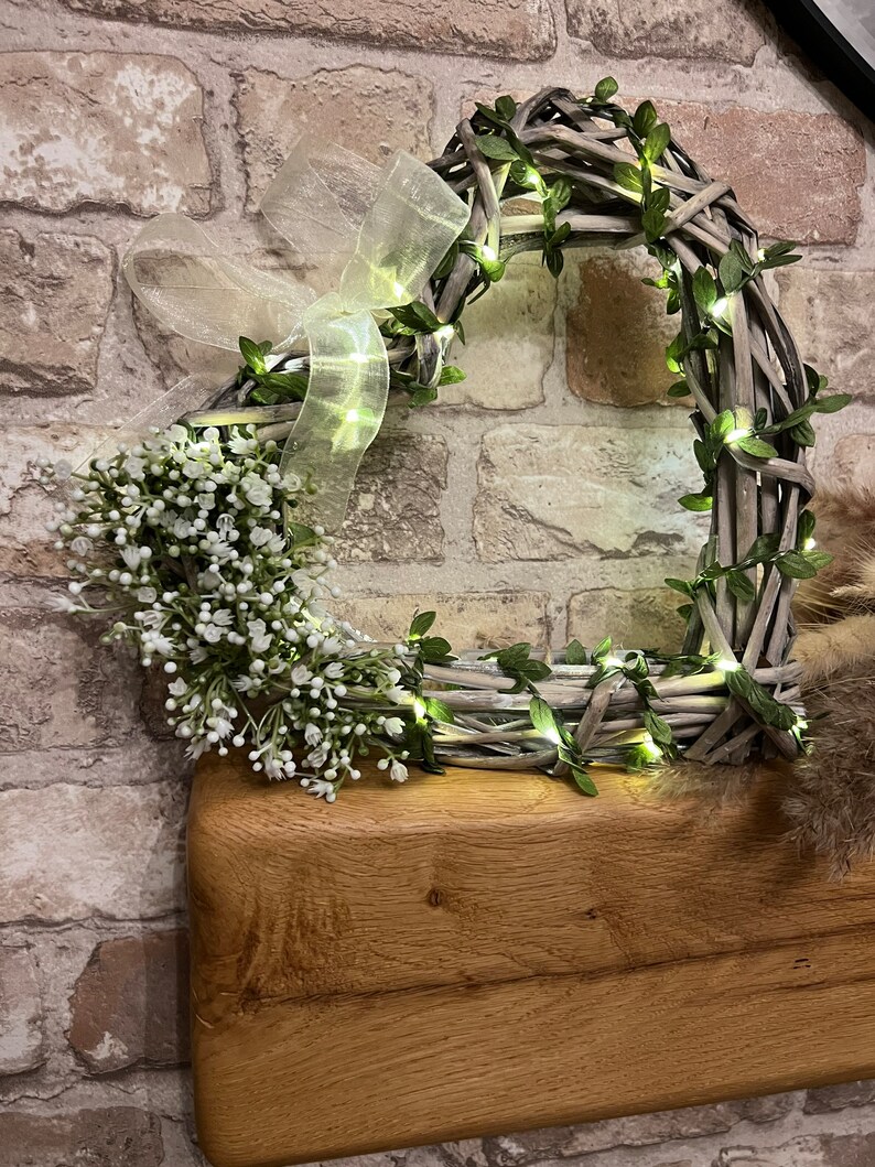 May include: A heart-shaped wreath made of woven twigs and greenery with white fairy lights wrapped around it. The wreath is decorated with a white sheer ribbon and a cluster of small white flowers.