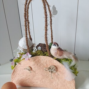 Könnte beinhalten: Ein pfirsichfarbener Papiermaché-Korb mit einem Seilgriff, verziert mit Moos, einem kleinen Vogel, einem weißen Ei und einem weißen Schmetterling. Der Korb steht auf einer weißen Oberfläche.