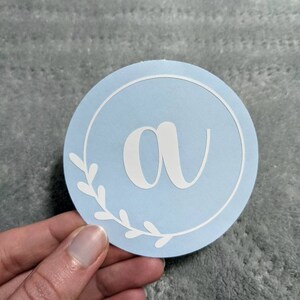 Op de afbeelding: Een ronde, lichtblauwe sticker met een witte cirkel rond de rand. De letter "a" staat in het midden van de cirkel in wit. Er zijn witte bladeren rond de cirkel.