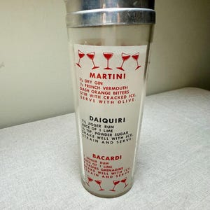 May include: A vintage cocktail shaker with a silver lid and a clear glass body. The glass features red text and illustrations of cocktail glasses, with recipes for Martini, Daiquiri, and Bacardi cocktails.