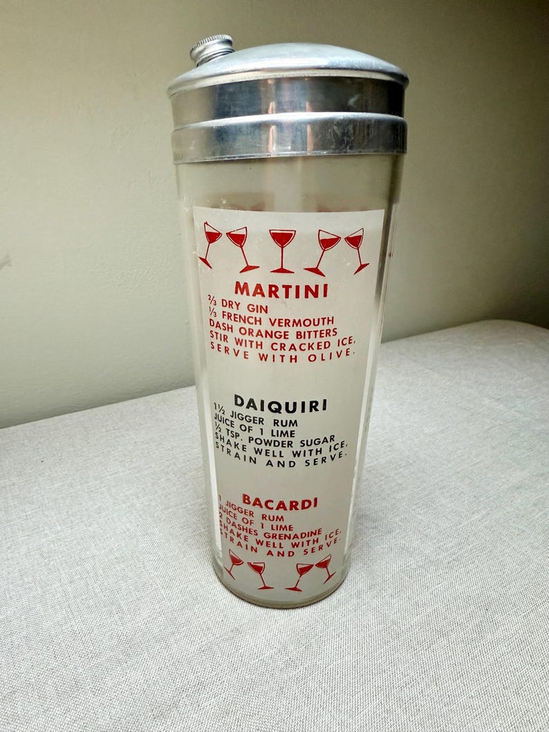 May include: A vintage cocktail shaker with a silver lid and a clear glass body. The glass features red text and illustrations of cocktail glasses, with recipes for Martini, Daiquiri, and Bacardi cocktails.