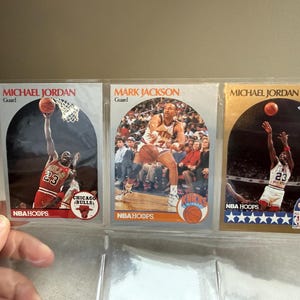 May include: Three basketball trading cards featuring Michael Jordan and Mark Jackson. The cards are in a clear plastic holder. The cards have images of the players in action, with the NBA Hoops logo and team names.
