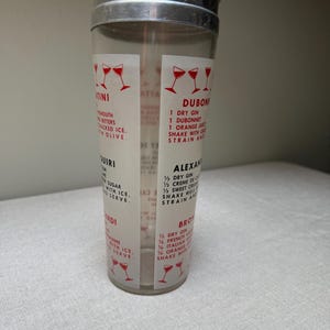 May include: A vintage cocktail shaker with a stainless steel lid and a clear glass body. The glass features red graphics of cocktail glasses and cocktail recipes. The shaker is designed for mixing drinks.