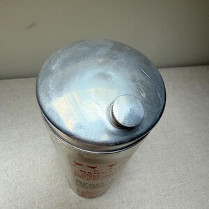 May include: A silver-colored cocktail shaker with a rounded top and a small, screw-on cap. The cylindrical body has red text and graphics. The shaker is designed for mixing drinks.