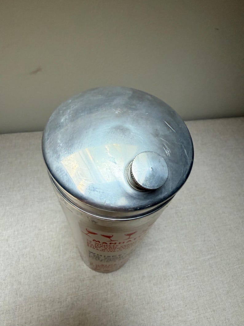 May include: A silver-colored cocktail shaker with a rounded top and a small, screw-on cap. The cylindrical body has red text and graphics. The shaker is designed for mixing drinks.
