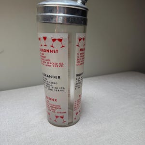 May include: A vintage cocktail shaker with a clear glass body and a silver metal top. The shaker features printed recipes for classic cocktails like Dubonnet, Alexander, and Bronx. Red wine glass illustrations accent the text.