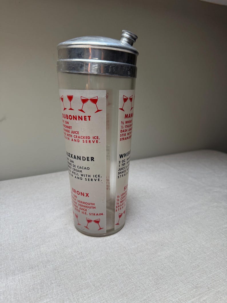May include: A vintage cocktail shaker with a clear glass body and a silver metal top. The shaker features printed recipes for classic cocktails like Dubonnet, Alexander, and Bronx. Red wine glass illustrations accent the text.