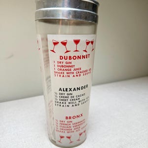 May include: A vintage cocktail shaker with a silver lid and a clear glass body. The shaker features printed recipes for classic cocktails like Dubonnet, Alexander, and Bronx. Red illustrations of cocktail glasses are also printed on the glass.