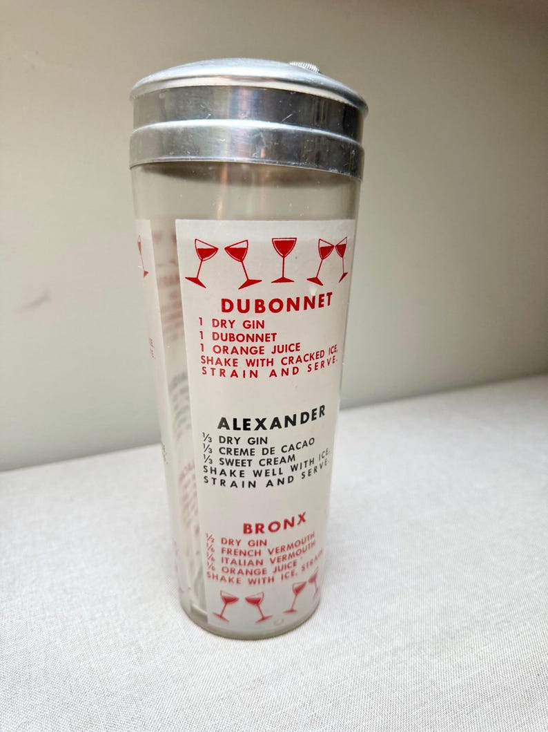 May include: A vintage cocktail shaker with a silver lid and a clear glass body. The shaker features printed recipes for classic cocktails like Dubonnet, Alexander, and Bronx. Red illustrations of cocktail glasses are also printed on the glass.