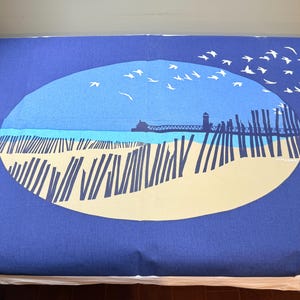 Könnte beinhalten: Ein rechteckiger blauer Wandbehang aus Stoff mit einer großen ovalen Grafik in der Mitte. Das Design zeigt einen Sandstrand mit dunkelblauen Holzzaunpfählen, einen Steg mit einem Leuchtturm und einen Schwarm weißer Vögel am hellblauen Himmel.