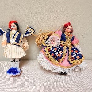 May include: Two traditional dolls. One doll is dressed in a blue and white outfit with a red hat. The other doll is wearing a pink and blue dress with a red hat and lace trim. Both dolls have painted faces.