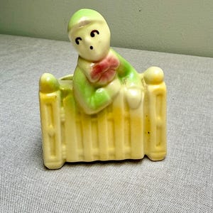 May include: A vintage ceramic planter featuring a figure in a green outfit leaning over a yellow fence. The figure has a light complexion, black eyes, and a red bow. The planter is a light yellow color.