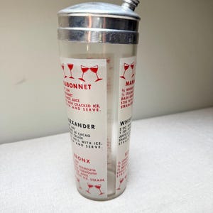 May include: A vintage cocktail shaker with a stainless steel top and a clear glass body. The glass features printed recipes for classic cocktails like Dubonnet and Alexander, with red graphic illustrations.